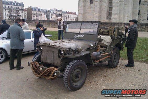 Olive drab military jeep with 'Charlotte' on windshield, parked next to a modern white car. People stand around