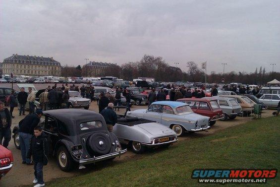 Outdoor gathering of people looking at rows of vintage cars parked on grass, with a large building in the background