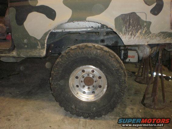 Off-road vehicle's rear wheel and fender with camouflage paint. A white box with red lettering is visible behind the wheel