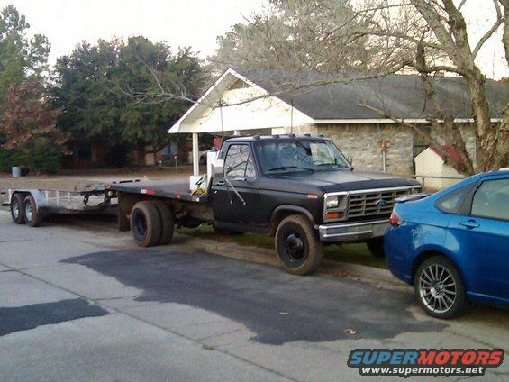 Black Ford F-350 flatbed truck with dual rear wheels, towing a trailer, parked next to a blue car