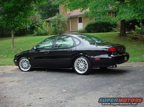 Black sedan parked on a street in front of a house. It has aftermarket wheels