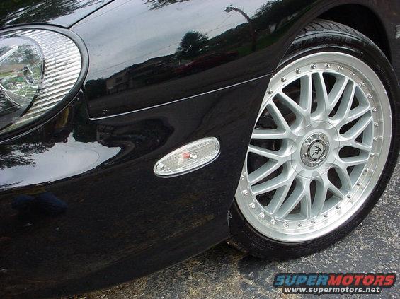 Black car front fender, clear turn signal, and silver aftermarket wheel. Supermotors.net logo visible