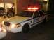 2002 Ford Crown Victoria - A white 2002 Ford Crown Victoria police car with its emergency lights activated is parked in front of a garage.