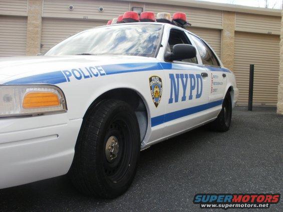 White 2002 Ford Crown Victoria police car with NYPD markings and light bar, parked in front of garage doors