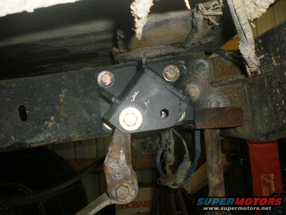 Close-up of a black metal bracket bolted to a rusty vehicle frame, with suspension parts and wiring visible
