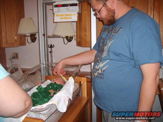 Newborn baby in green pajamas in hospital bassinet, with a man and another person looking on