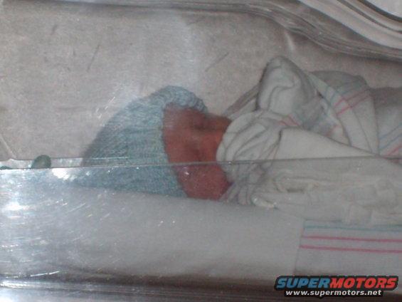 Newborn baby wearing a blue cap and wrapped in a striped blanket inside a clear incubator