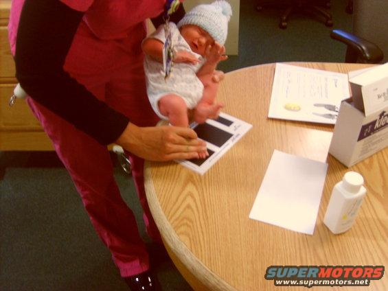 Newborn baby in a blue hat and onesie is held over a scale on a wooden table. Documents and a bottle of baby powder are nearby