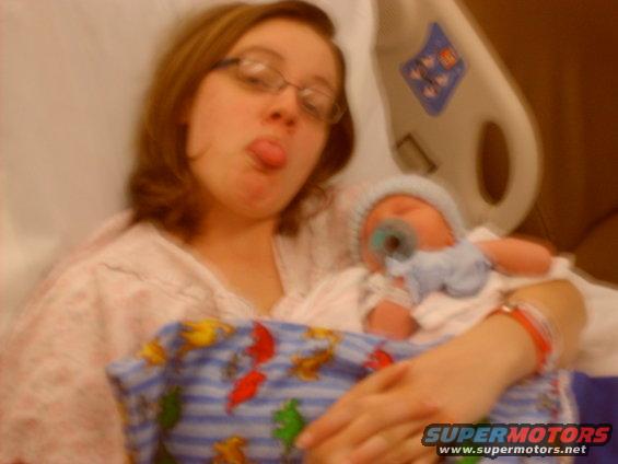 Woman in hospital gown sticking her tongue out while holding a newborn baby with a pacifier