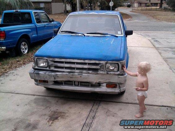 Toddler touching the headlight of a blue pickup truck. Another blue pickup is parked behind it