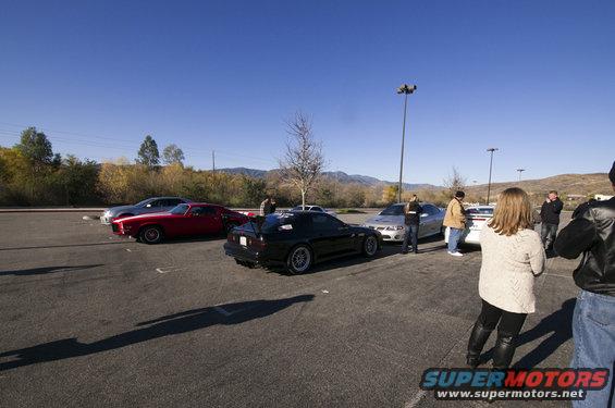 Black sports car with spoiler parked in a lot with other vehicles and people