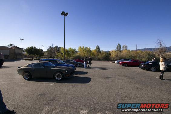 Several classic cars parked in a lot, including a dark green coupe in the foreground and a red sports car further back