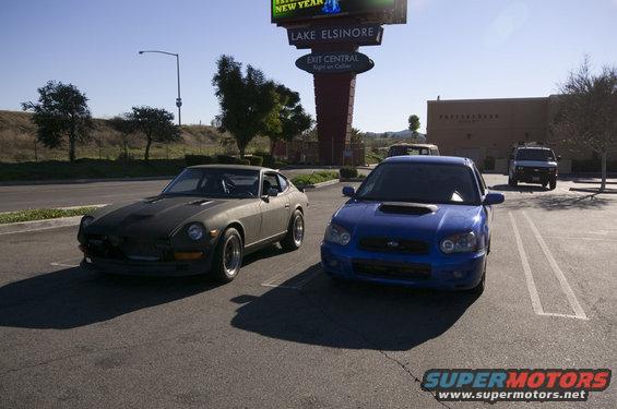Gray Datsun 240Z and blue Subaru WRX parked in a lot with a large sign in the background