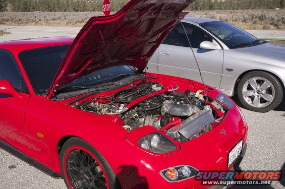 Red sports car with hood open, showing engine and aftermarket intercooler. Black wheels with red trim. Silver sedan behind