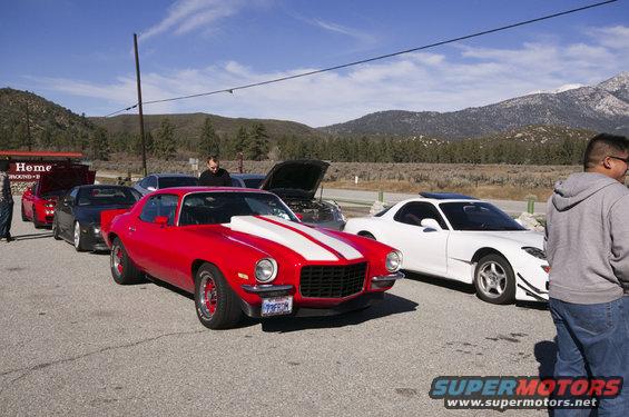 Red 1970s muscle car with white stripes, front 3/4 view. License plate visible