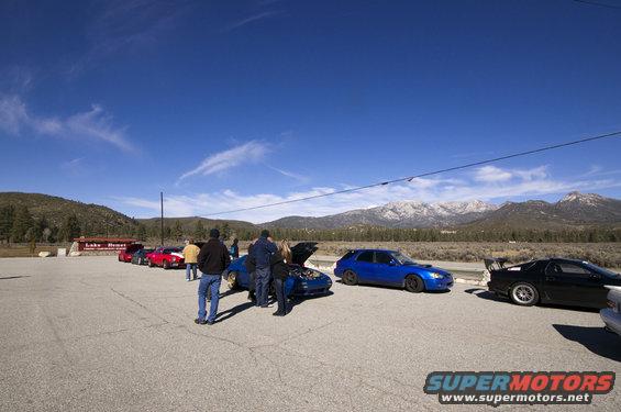 Several cars parked in a lot with people gathered around, mountains in the background