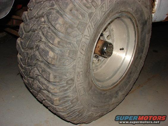 Close-up of a large off-road tire with aggressive tread. The wheel is silver with visible lug nuts. Part of the vehicle's frame is visible above the tire