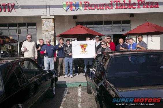 Group of guys holding a USMC banner in front of Round Table Pizza, with two black cars parked nearby