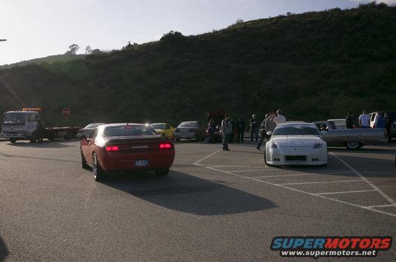 Red sports car from rear, parked in lot with other vehicles and people