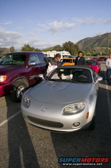 Silver Mazda MX-5 Miata convertible with a black soft top, parked in a lot with a red truck and other cars. The front grille and headlights are visible