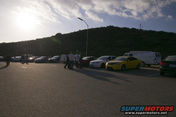 Parking lot scene with a yellow coupe, white sedan, dark sedan, and a white van. Several people are standing around