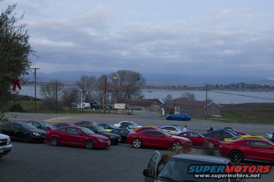 Cars parked in a lot with a lake and hills in the distance under a cloudy sky