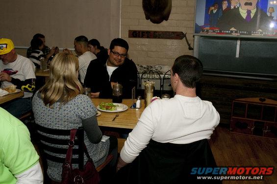 People sitting at tables in a restaurant, some eating, others talking. A TV is on in the background