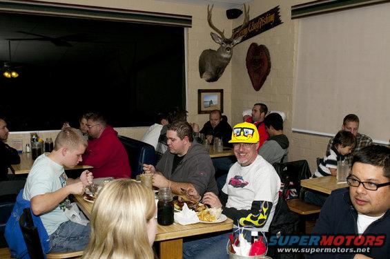 Group of people eating at tables inside a restaurant. A deer head is mounted on the wall
