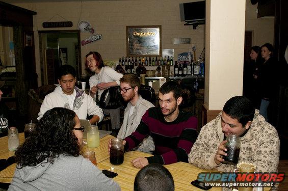 Group of people sitting around tables in a bar, with bottles on shelves behind them