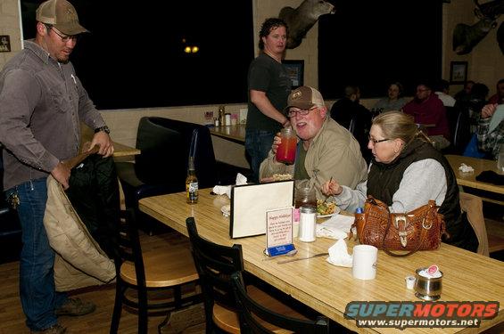 Man in cap pulls jacket from bag at table, guy drinks from jar, woman eats