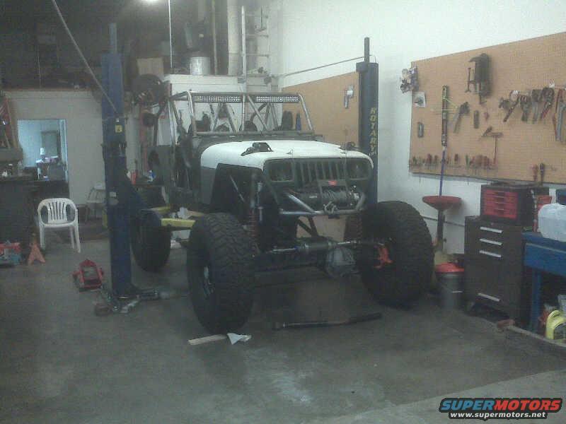 Jeep Wrangler on lift, white hood, gray body, large tires, engine exposed, tools on wall