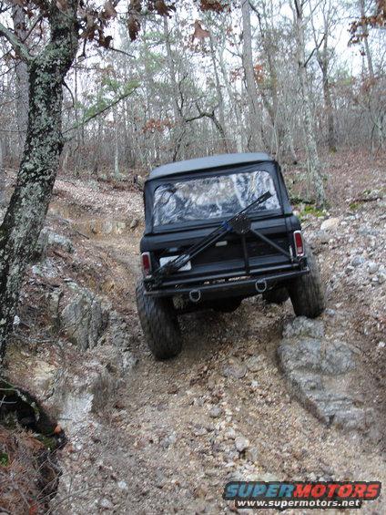 Black SUV with large tires navigating a rocky, wooded trail from the rear