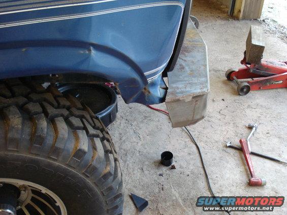 Close-up of a blue truck's rear fender and large tire, showing damage and a rusty aftermarket bumper. Tools and a jack are nearby