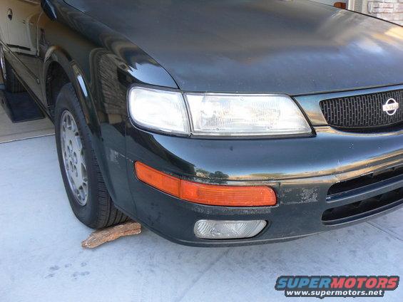 Front corner of a dark sedan with a Nissan badge on the grille. A piece of wood is under the front tire