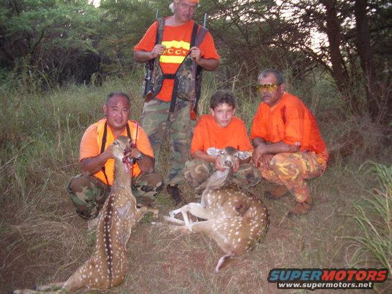 Four men and a boy in orange shirts and camouflage pants pose with two dead deer in a grassy, wooded area