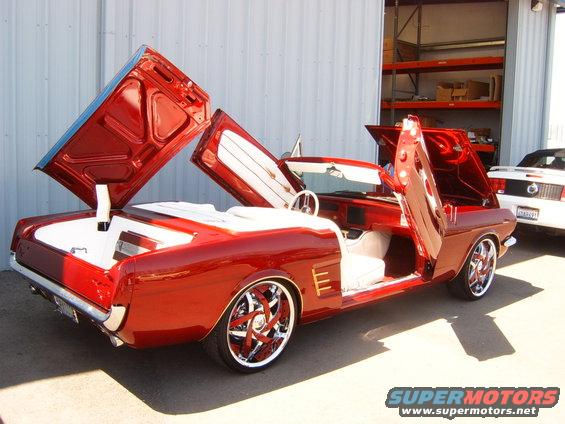Red convertible with doors and hood open, white interior, custom wheels. Another car is parked behind it