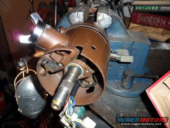 Brown steering column assembly held in a blue vise, with wires and a metal lever visible