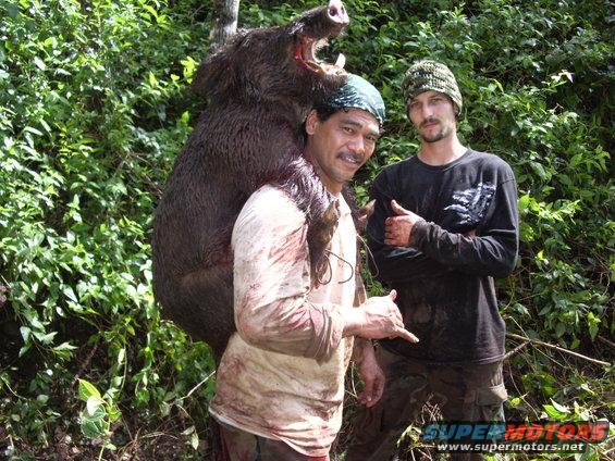 Two men carrying a large wild boar, with a website watermark at the bottom right