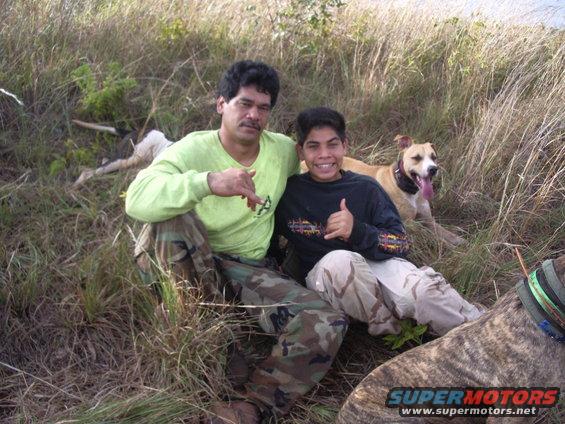 Two men and two dogs sit in dry grass, one man wears a lime green shirt and camouflage pants, the other a black shirt with a graphic design