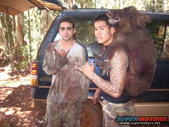 Two guys stand by a dark SUV in the woods, one carrying a large wild boar over his shoulder