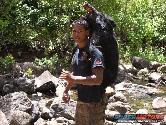Man with tattoos carries a large wild boar over his shoulders in a rocky, wooded area near a stream