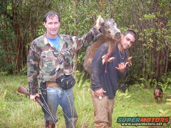 Two men pose with a wild boar carcass in a wooded area. A dog stands in the background