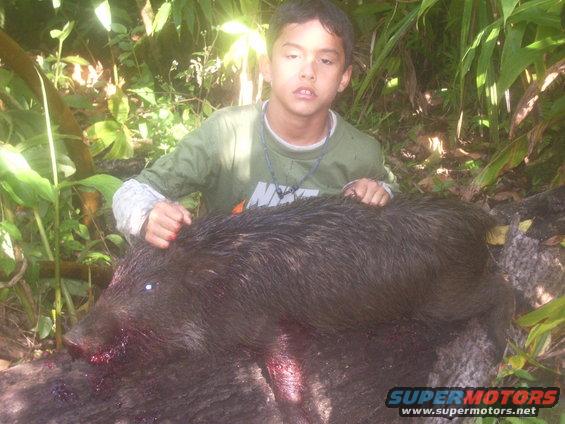 kid posing with a large wild pig he hunted, outdoors in foliage