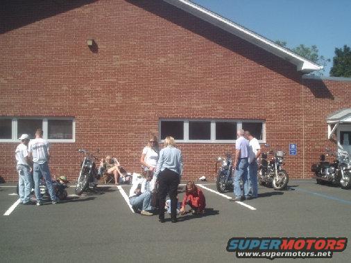 Several motorcycles parked in a lot with people standing and sitting around