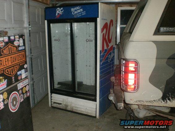 Rear of a light-colored vehicle with camouflage pattern and a red taillight next to an RC Cola refrigerator