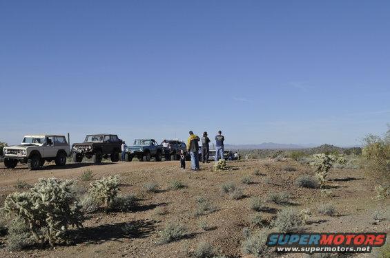 Several off-road vehicles parked on a dirt road in a desert landscape with people standing around