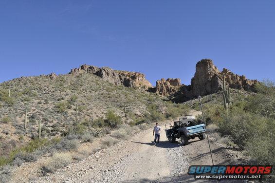 Two off-road vehicles on a dirt road in a desert landscape with rocky hills and cacti under a clear blue sky