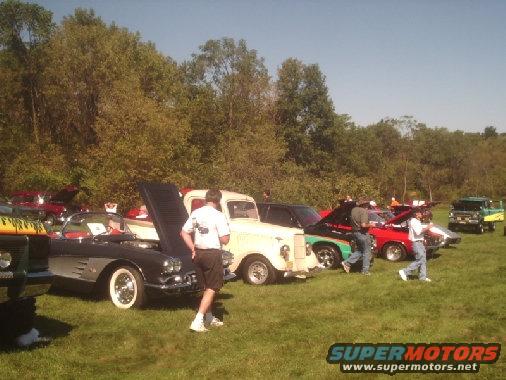 Cars parked on grass at a show. Some hoods are up. People are milling around