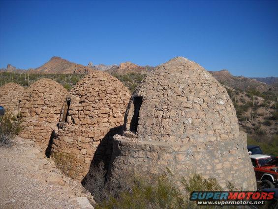 Stone kilns in a desert with mountains in the background. Two vehicles are partially visible on the right