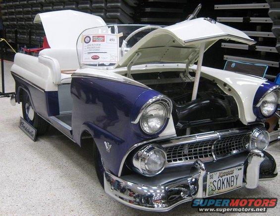 Custom golf cart resembling a 1955 Ford Fairlane, white and blue, hood open, Kansas license plate SQKNBY